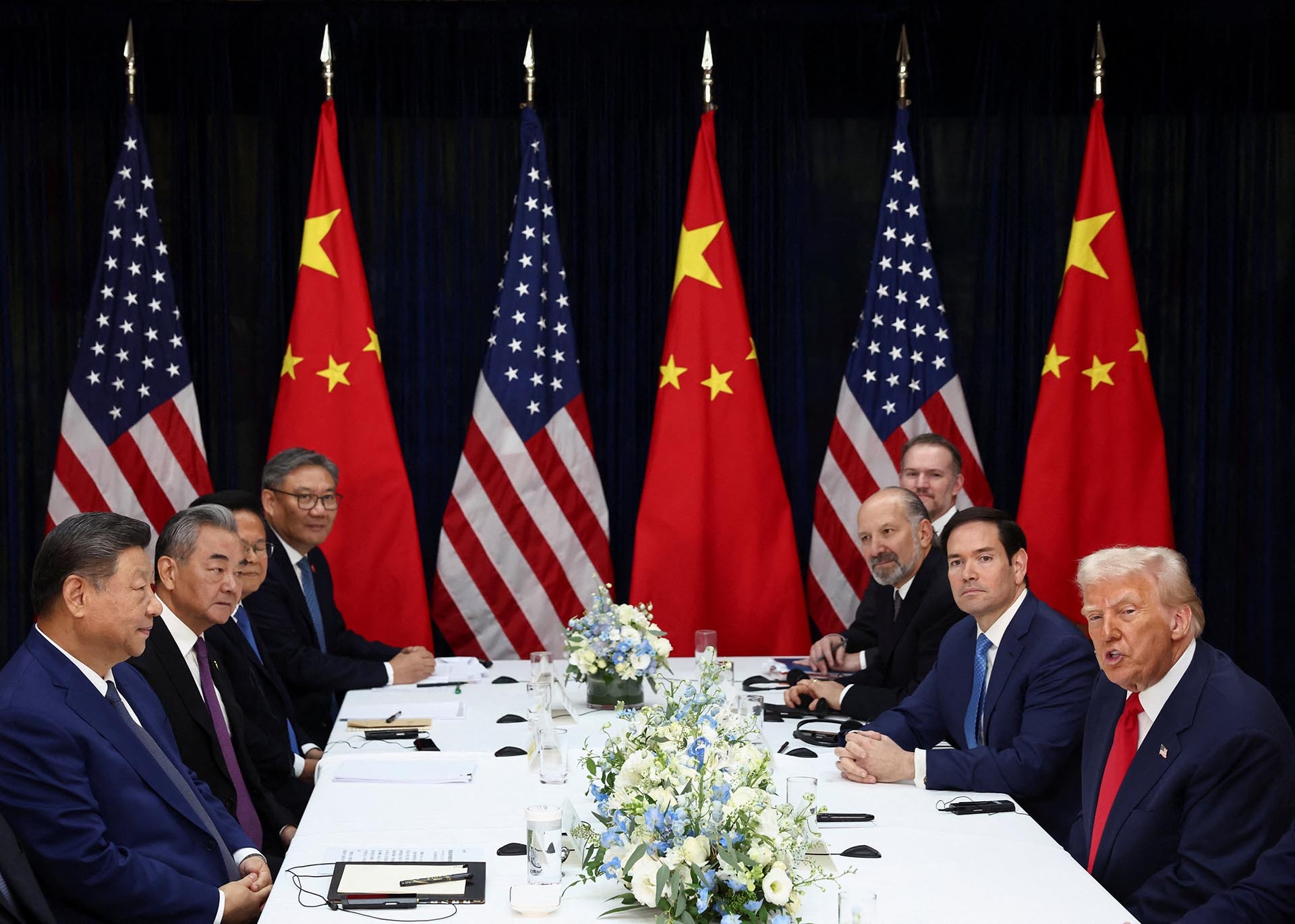 President Trump and President Xi sit across from the table from each other. Other men sit beside them. Chinese and American flags stand in the background