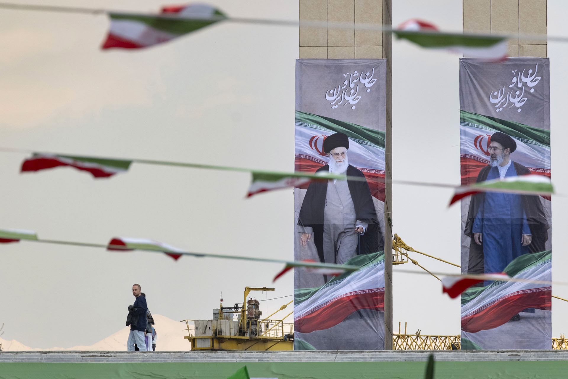 An Iranian man walks past the portraits of late Supreme Leader Ayatollah Ali Khamenei and the new Supreme Leader Ayatollah Mojtaba Khamenei during a flag ceremony marking Iran's Islamic Republic National Day in the Abbasabad Cultural and Tourist Area in central Tehran, on April 1, 2026.