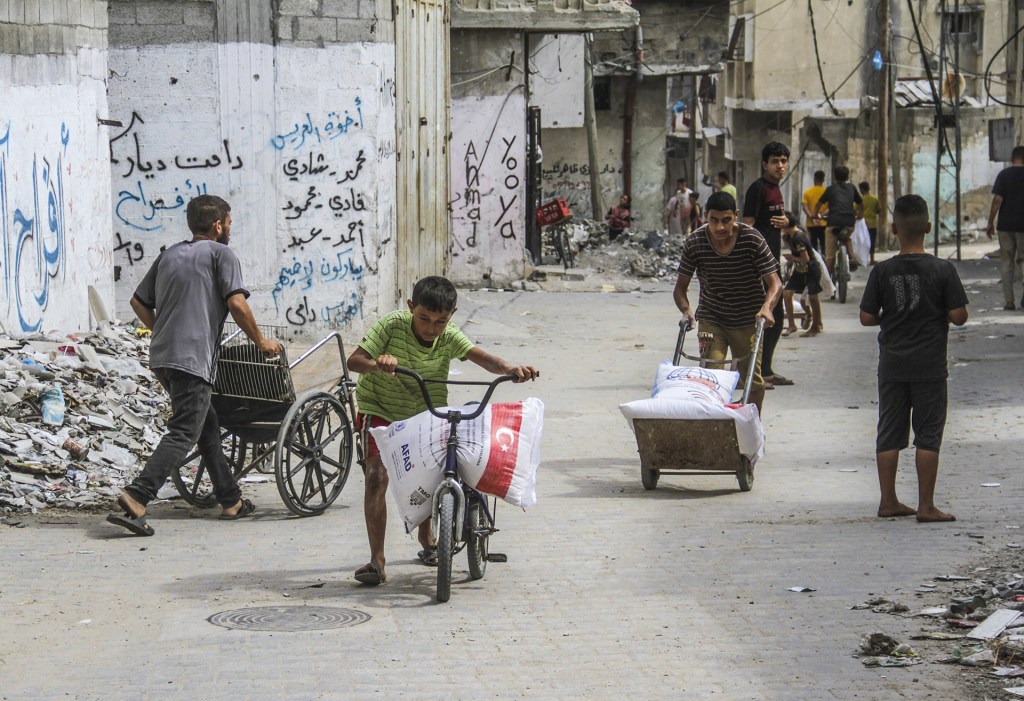 A young boy wheels a bike carrying a bag of food aid along a street, with others following him back to their camp.